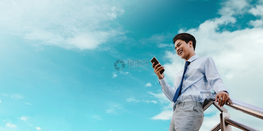 在现代人云蓝色天空的城市生活方式中使用移动电话作为背景低角度视的年轻人笑快乐的年轻商人肖像现代人云蓝天的模型常设图片
