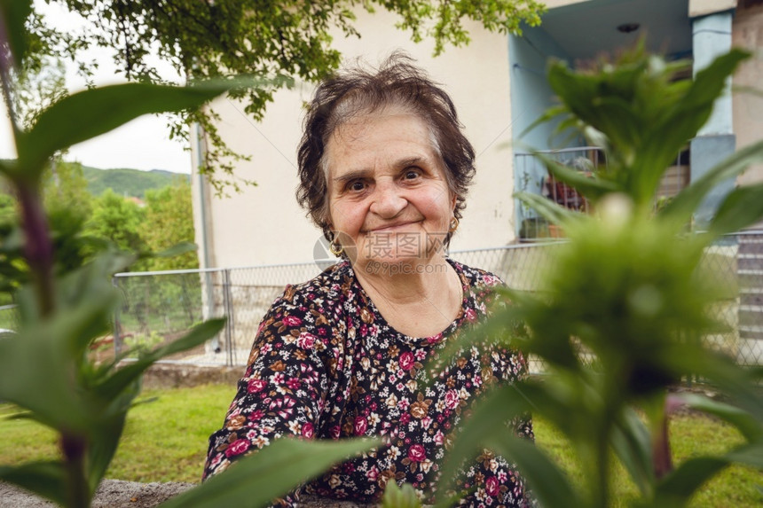 乡村的站在农庄她家中的房屋前坐在子面的老退休者caucasiansian女农民肖像看着绿色植物之间快乐真实人微笑的镜头观看着摄影图片