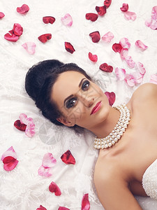 黑发穿着婚纱的美丽新娘年轻女子躺在床上带着玫瑰花瓣化妆品图片