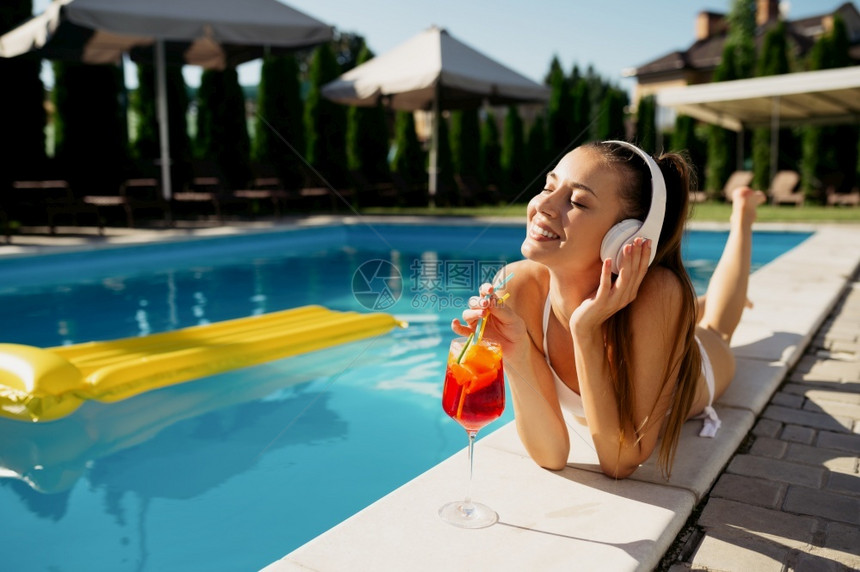 游泳的在池边缘听音乐和喝汽水的妇女耳机在游泳池边缘听到音乐和喝小鸡尾酒的人欢乐快们在暑假泳池边户外的节日派对上玩得开心妇女休闲人图片