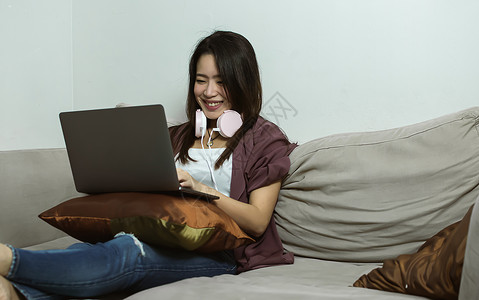 活的肖像人使用笔记本电脑和耳机的亚洲年轻美女在家用生活风格技术和新常识概念客厅休息时使用笔记本电脑和耳机背景图片