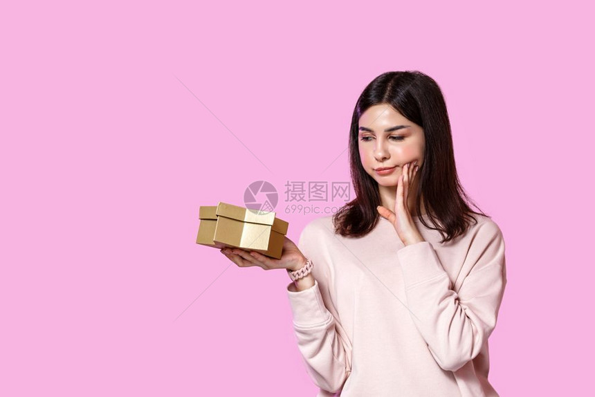 不满意一个年轻女孩穿着毛衣手里拿着一个礼物盒的不满和失望在粉红色孤立的背景上不喜欢伤心图片
