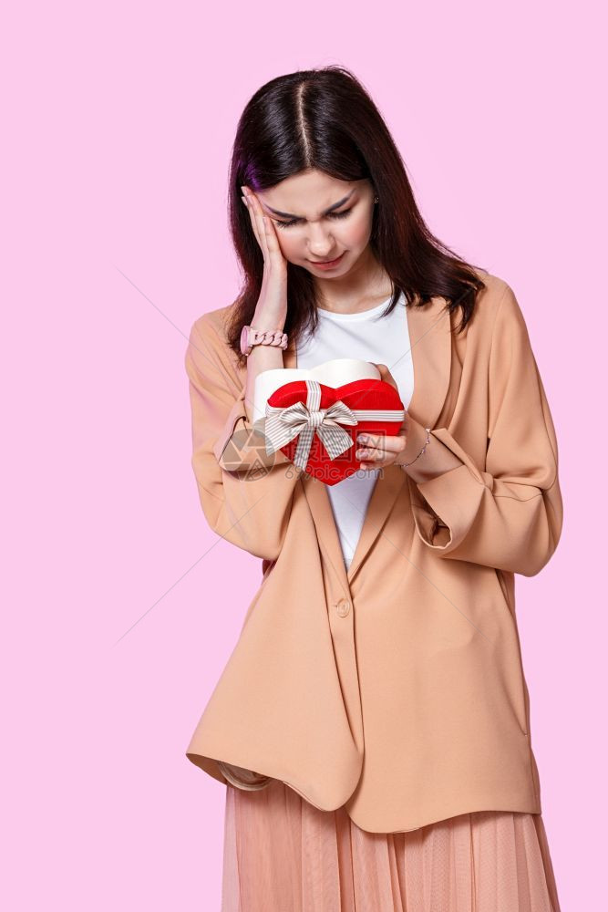 失望的女朋友周年纪念日一个轻女孩穿着一件外套手里拿着一个礼品盒的物对粉红色孤立的背景感到不满和失望图片
