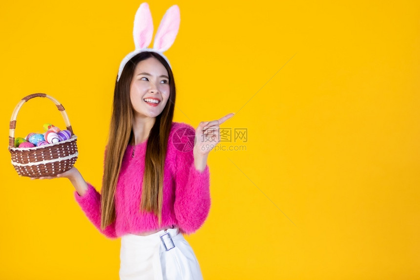 穿着兔子耳朵的亚洲快乐女青年手拿着一个篮子配彩色复活节鸡蛋在黄色空版间工作室背景上孤立无闻漂亮的空白庆典图片