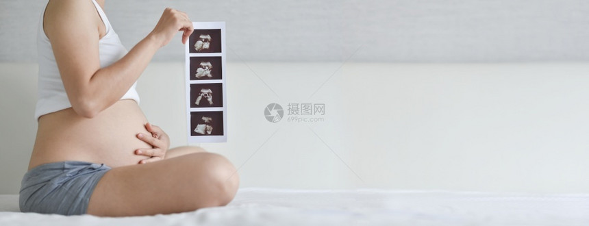 亚洲幸福孕妇坐在白间怀着超声波图像与未出生婴儿的声波X光在床上触摸肚子时坐在胸怀超声像的亚洲快乐孕妇Banner怀孕概念产前后护图片