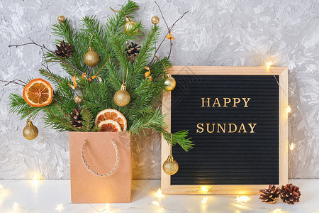 黑色星期天分支机构黑色信板上的快乐星期天文字和带有圣诞装饰的冷杉树枝节日花束在工艺包和桌子上的灯花环明信片模板概念你好冬天星期快乐的文字在背景