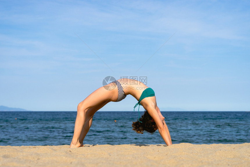 晴天自然阳光明媚的日子里海滩上的年轻女子在海边的桥位置SetuBandhasanaasana练习瑜伽身体图片