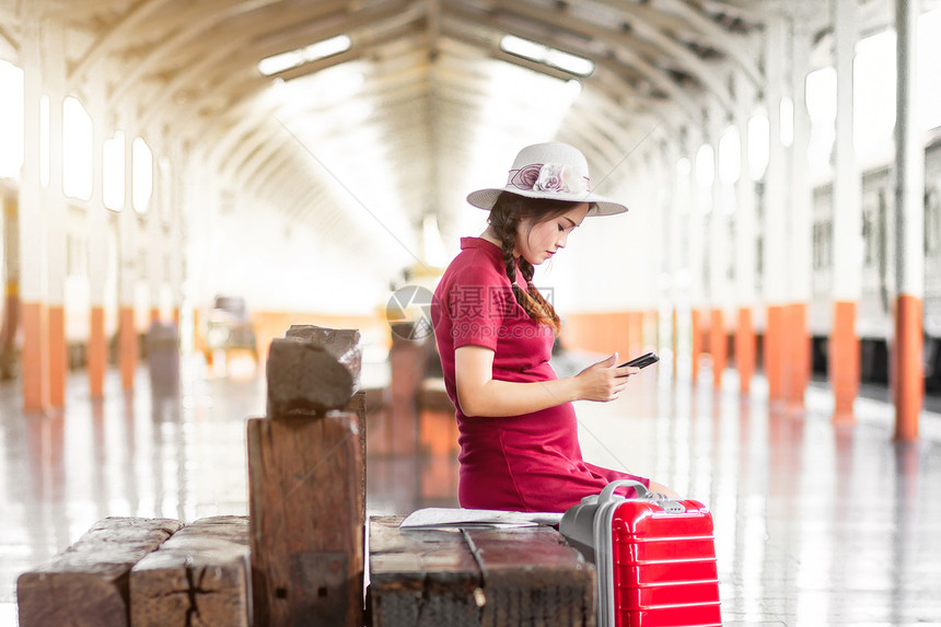 快乐的身着红色礼服携带行李的亚洲妇女怀孕在火车站旅行时看到带红色手提箱的SmartphoneSmartwhone夏天电话图片