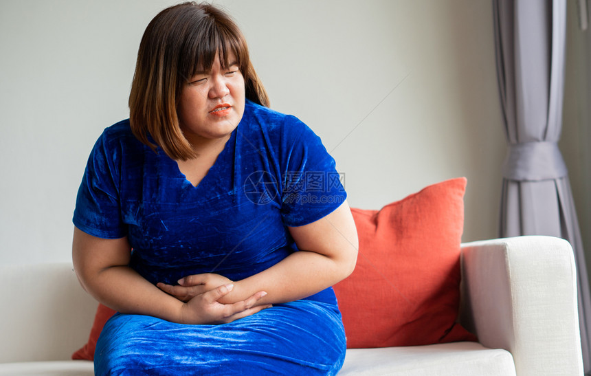 发牢骚痛便秘超重的亚洲妇女坐在客厅沙发上由于胃炎和保健概念手抓着肚子b由于胃部不适症和健康护理概念图片