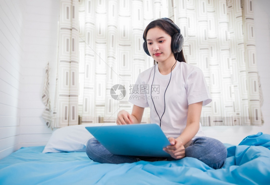 屋爱好亚洲快乐的年轻妇女戴耳机听音乐歌曲观看网站在家床上用笔记本电脑在网上购物校对Soup女图片