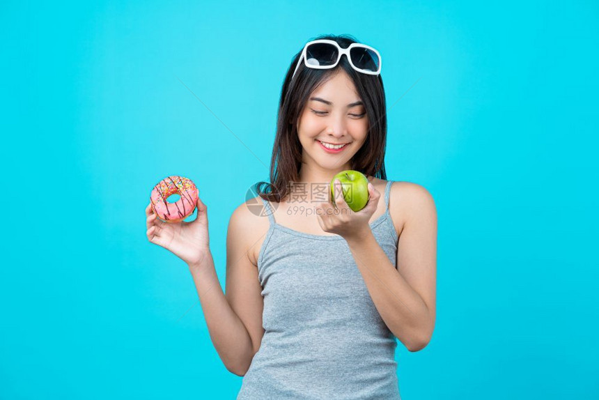 成人有吸引力的亚洲年轻妇女持有甜圈盘或绿苹果水并以孤立的蓝色背景体重下降和避免为饮食健康而吃垃圾食品的方式在甜圈盘或绿色苹果水之图片