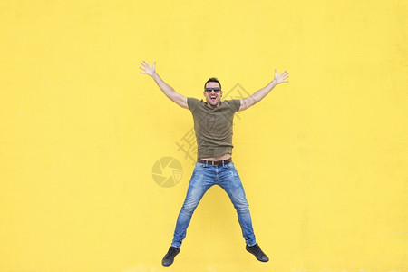 外部身戴太阳眼镜的年轻时尚男子在黄墙上跳跃牛仔裤黄色的图片
