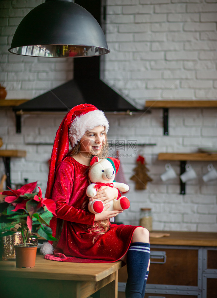 展示圣诞快乐戴着帽的可爱小女孩抱着一只泰迪熊坐在厨房里等待假期一个奇迹和满足愿望的时刻圣诞快乐戴着帽的可爱小女孩抱着一只泰迪熊坐图片