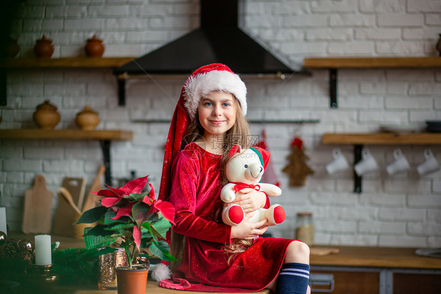 红色的一种庆典圣诞快乐戴着帽的可爱小女孩抱着一只泰迪熊坐在厨房里等待假期一个奇迹和满足愿望的时刻圣诞快乐戴着帽的可爱小女孩抱着一图片