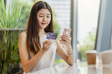 为了亚洲人使用带手机的信卡在咖啡店或办公场所在线购物的亚洲年轻妇女除了窗户镜子技术资金钱包和网上支付概念信用卡模型外还信卡和手机背景