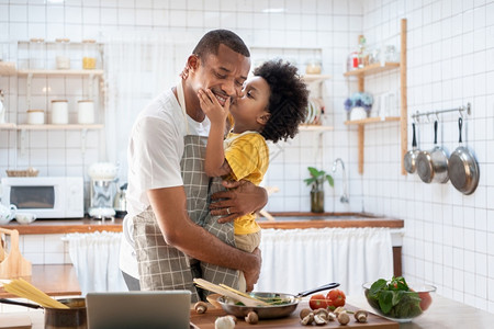 可爱的童年父母非洲可爱小男孩在黄色的闲散中和爸亲吻同时在家做饭快乐笑的非裔美国人父亲在厨房欢乐的黑人家庭爱情拥抱和着他的儿子背景图片