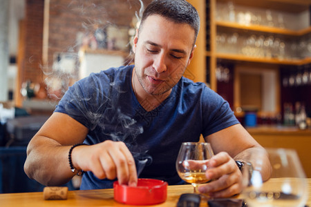 托盘闲暇坐在桌边着的年轻天主教男子的肖像在餐馆或家中拿着白兰地或杯子的家里扑灭了一根烟花抽到灰缸时穿着蓝色T恤衫的烟雾熄灭背景图片