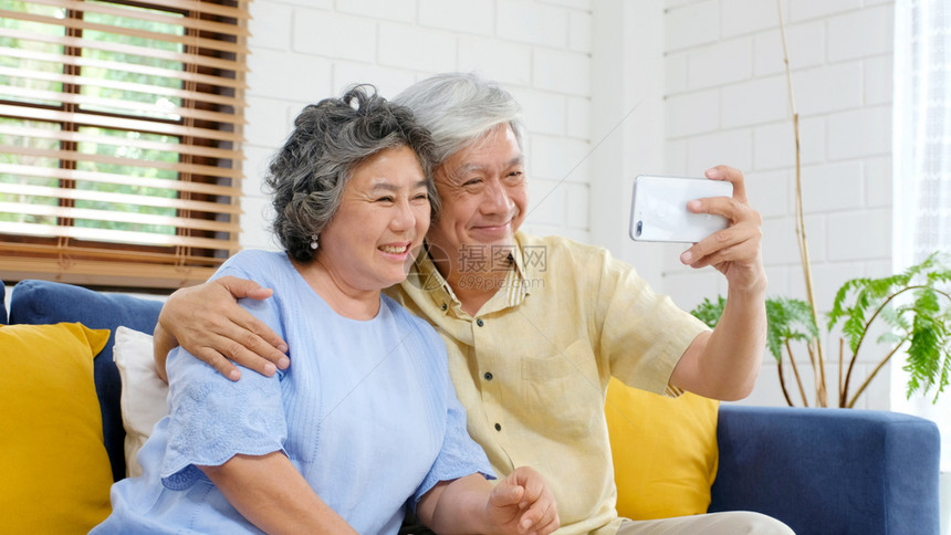 人们小工具电话快乐的老年亚裔夫妇在家里客厅自拍在欢乐时刻活跃的老年人有技术和生活方式的临时退休者以及具有技术和生活方式的临时退休图片