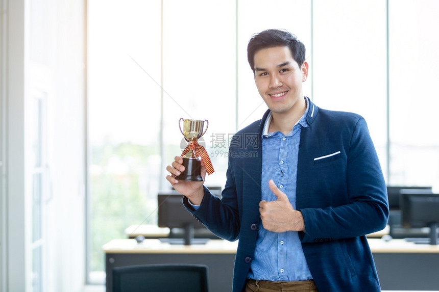 成功感觉的胜利者肖像尖叫着英俊的年轻亚洲商人拿着冠军杯在办公室背景中举起大拇指在办公室里杯子工作聪明的图片