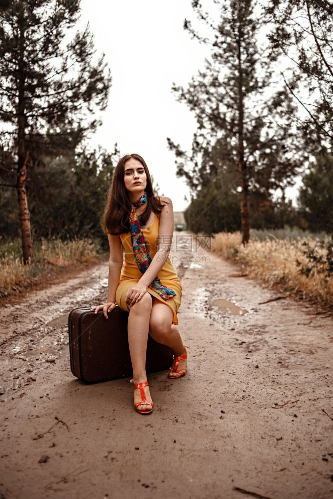 身着黄色裙子的年轻美女穿着彩色围巾坐在雨后湿泥路上的旧式手提箱吸引人的丰富多彩优雅图片