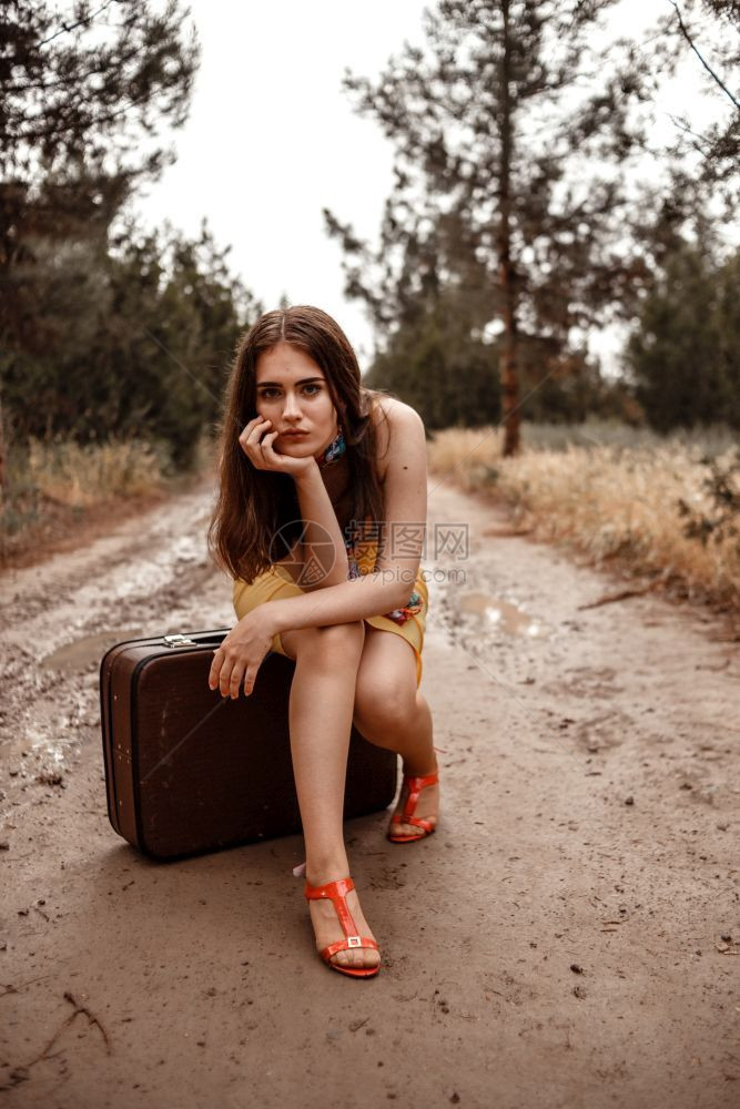 身着黄色裙子的年轻美女穿着彩色围巾坐在雨后湿泥路上的旧式手提箱复古的随意坐着图片