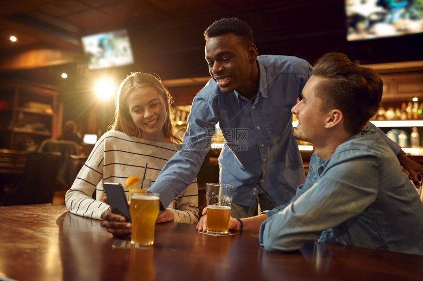 室内的年轻朋友们在电话上看照片在酒吧桌边玩乐一群人在酒吧休息晚上的生活方式友谊庆祝活动朋友们在酒吧里看手机的照片啤酒图片