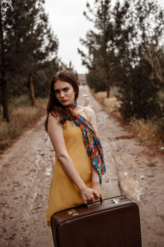 浪漫的白种人身着黄裙子的年轻美女围巾颜色多彩穿着旧式手提箱在雨后湿泥地路上户外图片