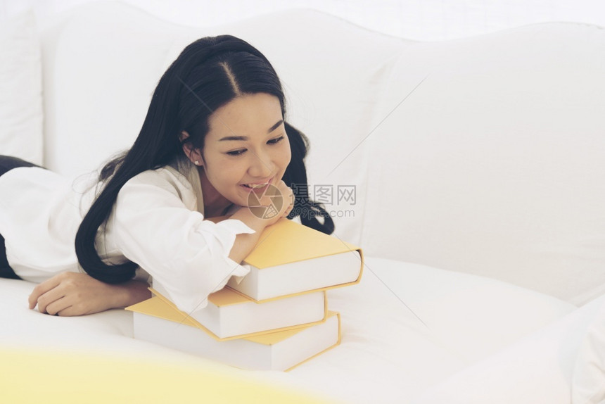 漂亮的难年轻亚洲女学生大在沙发上用手握着教科书的亚裔青年女子学生大与丰富多彩的枕头研究一起坐在沙发上很难获得更好的年级教育概念读图片
