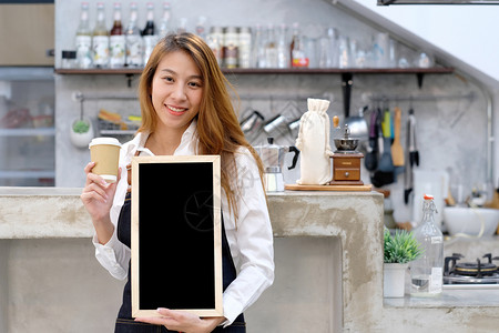 咖啡店黑板展示年轻的女亚洲咖啡师拿着一次咖啡杯和空黑纸板在咖啡馆的柜台背景食物和饮料概念上面带微笑的脸曼谷店铺背景