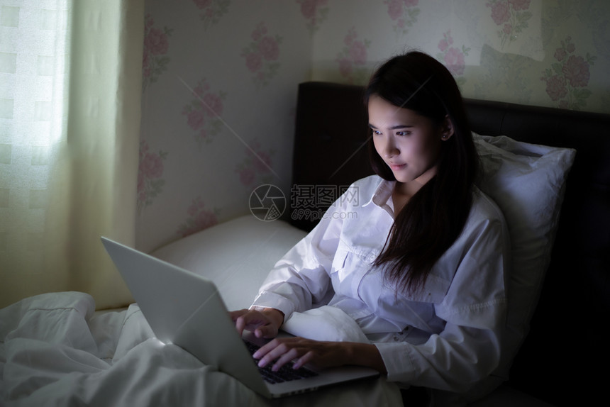 摄像头细胞年轻的亚裔妇女用笔记本在床上夜间工作她微笑并享受在家工作图片