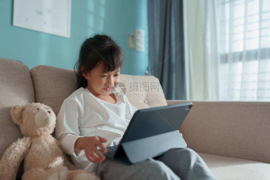 游戏快乐的亚洲小女孩在沙发上坐着时使用平板电脑早上在家和泰迪熊坐沙发上微笑娱乐图片