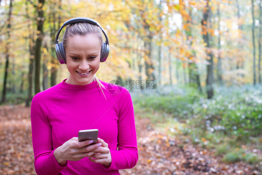锻炼身体流媒耳机妇女选择在秋天林地活动前通过移电话倾听音乐然后在秋天的森林进行锻炼图片