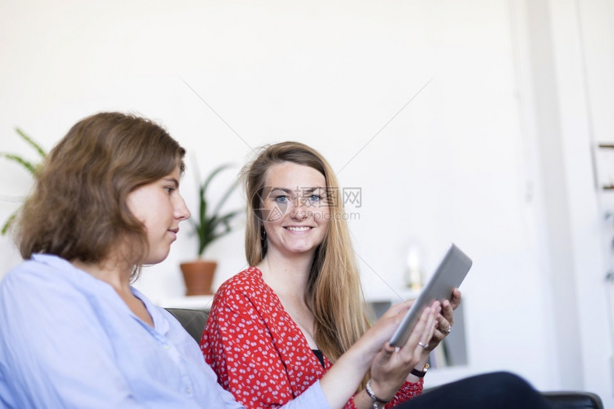 朋友们乐趣坐着在沙发上同时使用平板电脑和微笑的两名美丽年轻女青在家中头部照片图片