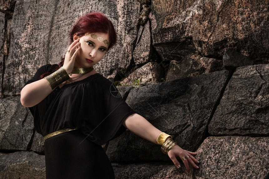 妖娆神秘头发美丽的化妆品女孩身着黑色外衣和古董珠宝背面有岩石墙图片