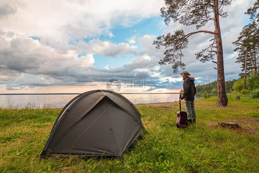 在湖岸露营帐篷附近站着吉他享受日落时风景的旅游者与自然支撑云靠近图片