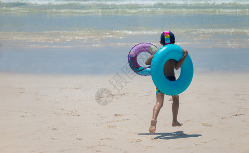 青年带着两个游泳环的男孩在海滩上欢快奔跑背部晴天图片
