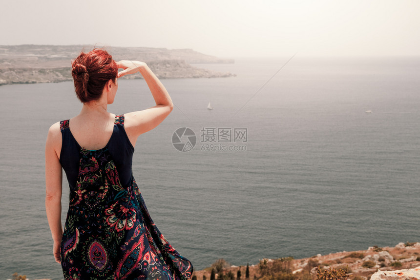 红发的年轻姑娘穿着长裙随风而动手从悬崖向上靠着头朝下从悬崖向海边看远乐趣强光白种人图片