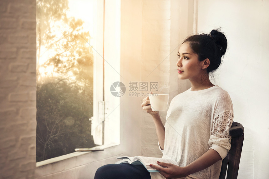 深思熟虑的年轻美女白布有书和咖啡杯坐在窗外看清晨荒凉的风景室内椅子美丽图片