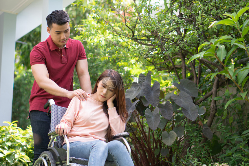疾病亚洲妇女坐在轮椅上亚洲妇女坐在轮椅上A男人站在轮椅后感到痛苦和正在鼓励他的妻子因为的脚意外事故而使腿部受伤无效的家庭图片