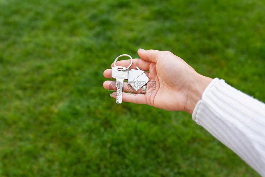 女士执照在绿草的背景下一个女孩手中的新房子或公寓钥匙链形式为金属房子的钥匙新或公寓手中的金属房子形式钥匙链一个以绿草为背景的女孩图片