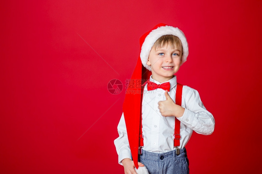 复制圣诞快乐可爱开朗的小男孩戴着红色背景的圣诞老人帽子一个快乐的童年有梦想和礼物庆典情感图片