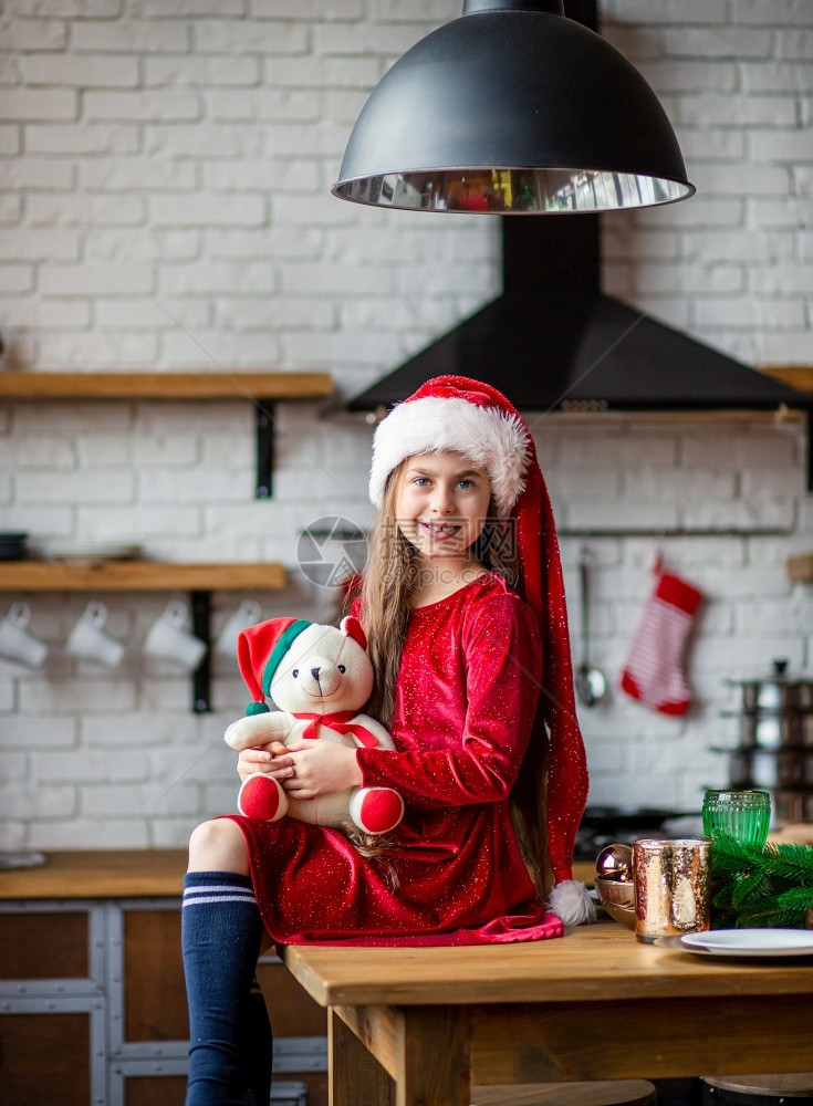 年轻的童展示圣诞快乐戴着帽的可爱小女孩抱着一只泰迪熊坐在厨房里等待假期一个奇迹和满足愿望的时刻圣诞快乐戴着帽的可爱小女孩抱着一只图片