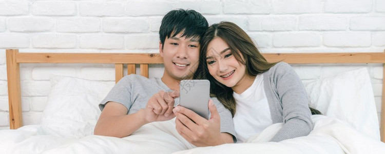 白种人妻子家卧室使用智能手机情侣和生活方式男朋友和女概念的亚洲快乐爱人现场报道在床上使用技术智能电话在家中卧室播放东南背景