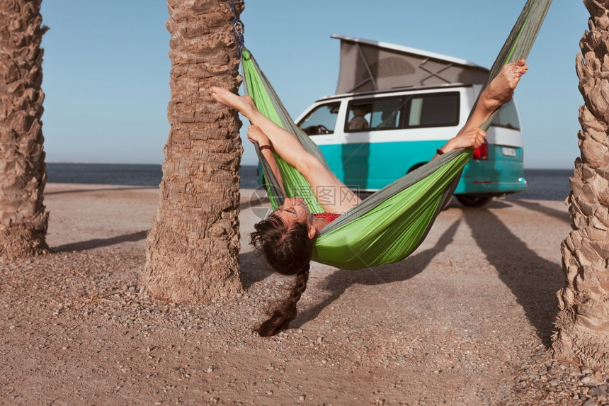 露营车妇女躺在沙滩上棕榈树之间的吊床上背着一辆野营车在海滩上搭起的吊床妇女躺在沙滩上棕榈树之间的吊床上假期人们图片