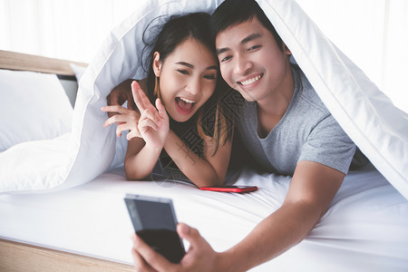亚洲情侣躺在家里玩手机和自拍的床上躺在游戏手机上聊天卧室请享用图片