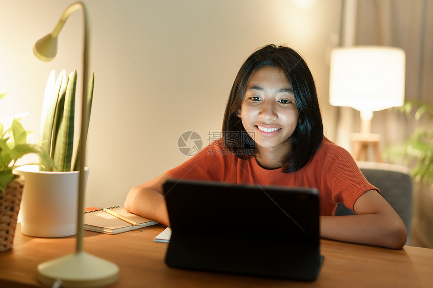 青年孩子药片家教做庭功课和在线学习的亚裔女孩在晚上坐书桌旁时与平板电脑上教师一起用数字平板电脑进行家庭教育做作业和网上学习图片