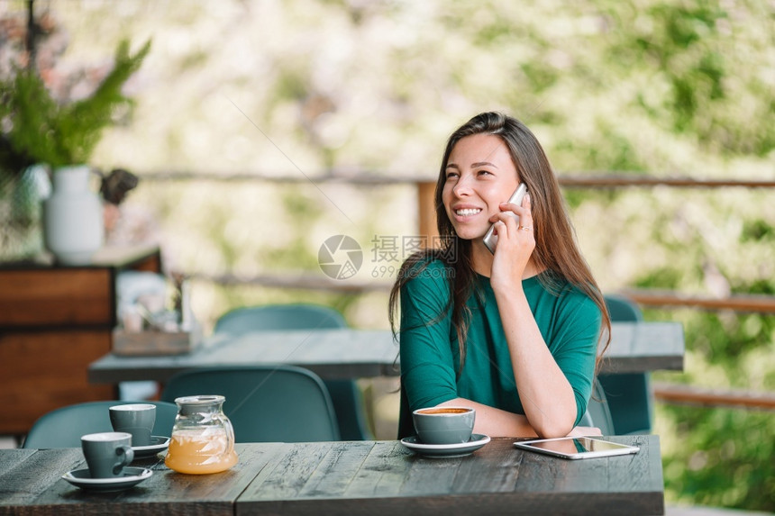 在业余时间单独坐在咖啡店的有智能电话年轻女子在户外咖啡馆打电话同时独自坐在咖啡店里青年杯子经过图片