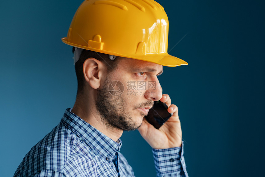 行业在蓝墙前穿着黄色保护安全头盔和衬衫的年轻男子工程师近距离肖像隔着智能手机在电话交谈边视线上进行称呼沟通图片
