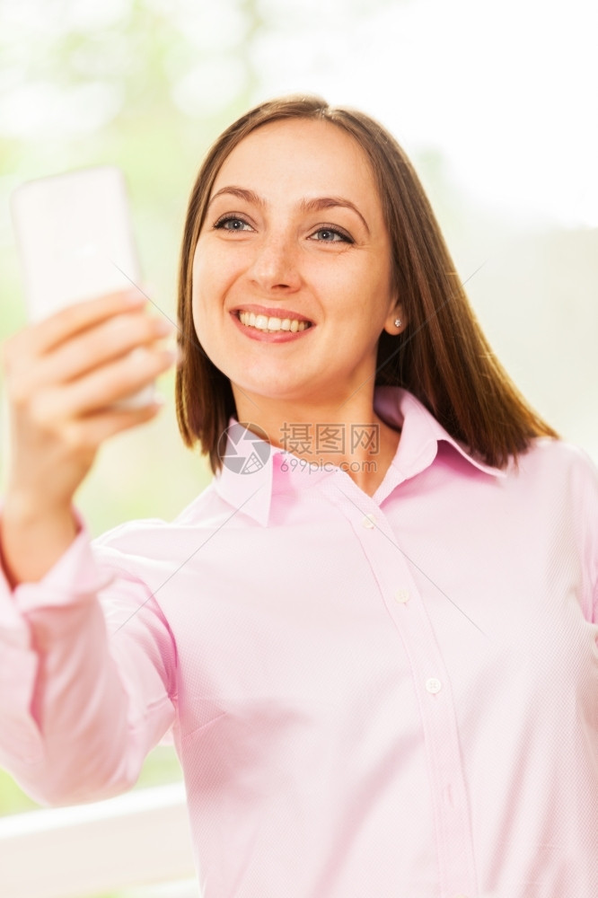 摆姿势随意的带着粉红衬衫笑脸caucasians女人正在用她的手机拍自头发图片
