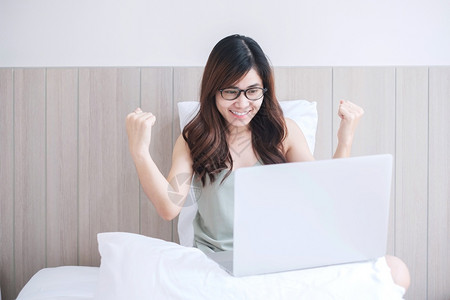 快乐的亚洲女坐在白床上并使用电脑笔记本年轻成女在家工作或网上购物时微笑和欢乐的年轻成女自由者生活方式和技术概念年轻的情感支付搜索高清图片素材